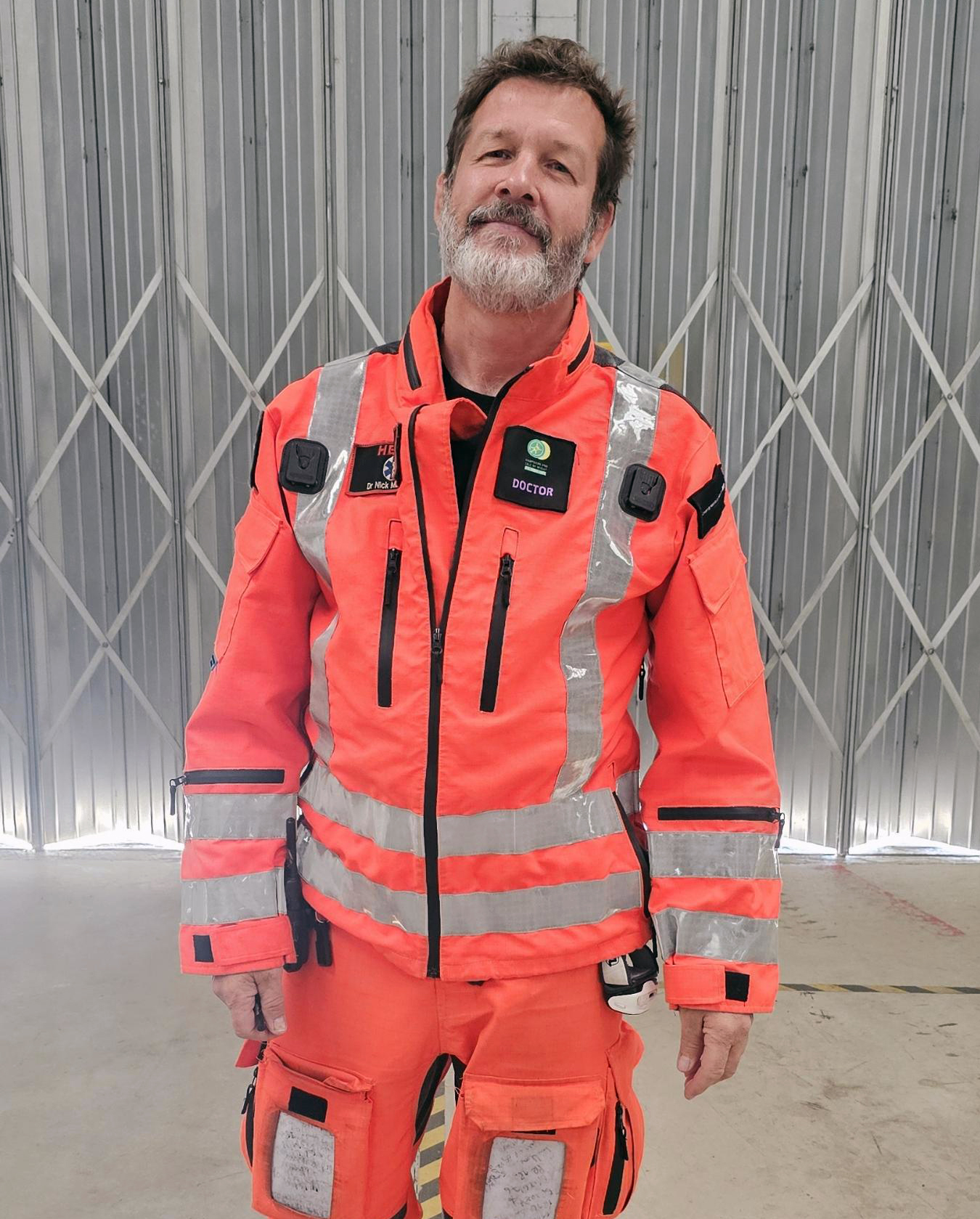 Dr Nick Maskery is wearing a full flight suit and is standing in the airbase hangar. The grey metal doors are behind him and he is smiling to camera.