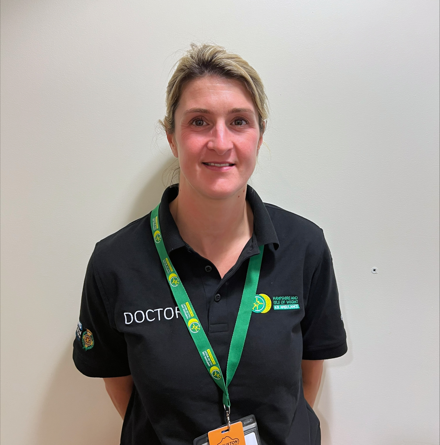 Dr Anna Barrow is wearing the doctor polo shirt and charity lanyard. She is standing in front of a cream wall and smiling to the camera.