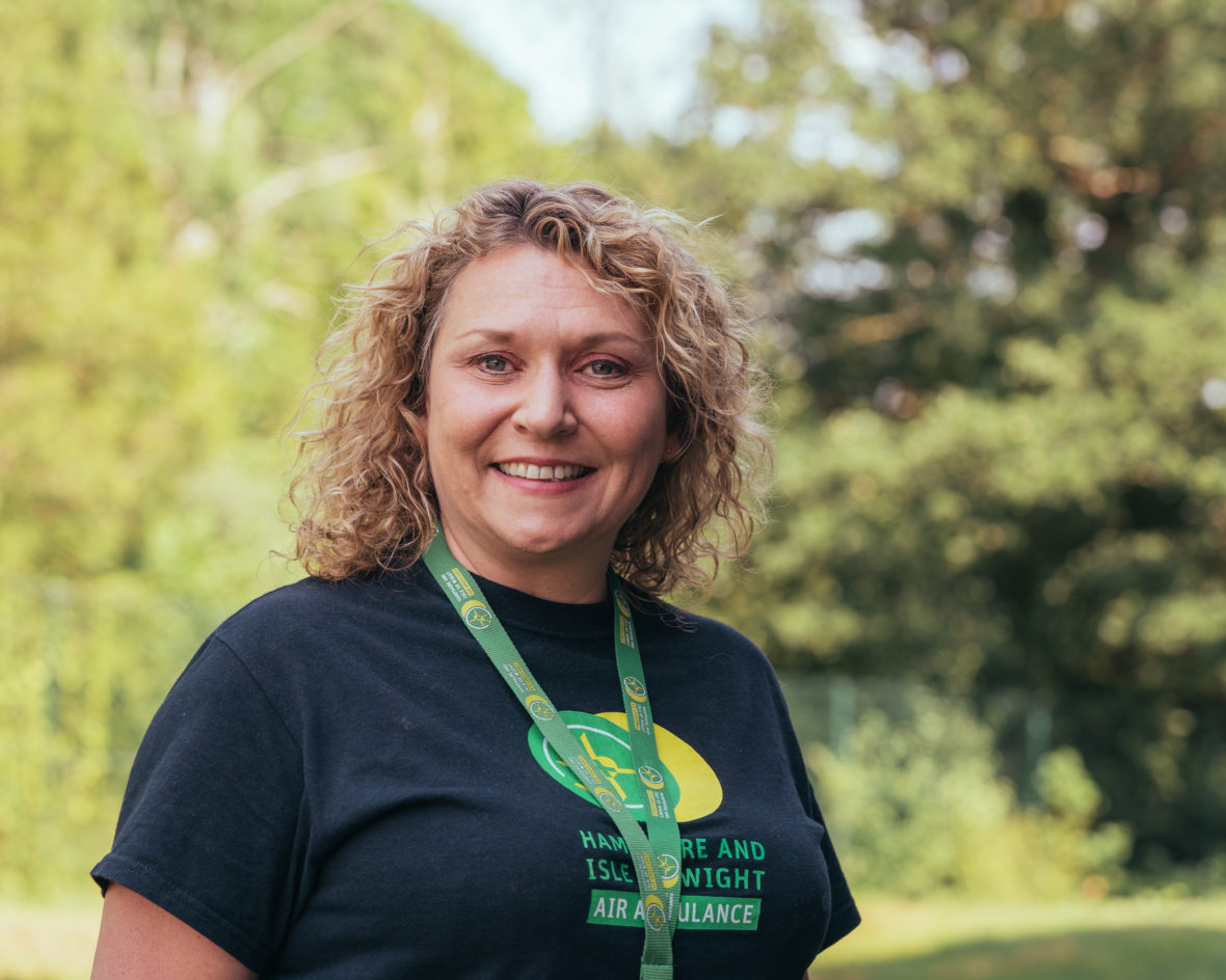 Nikki Harris is smiling to camera. She is wearing a charity t-shirt and lanyard. She is stood outside on a sunny day.