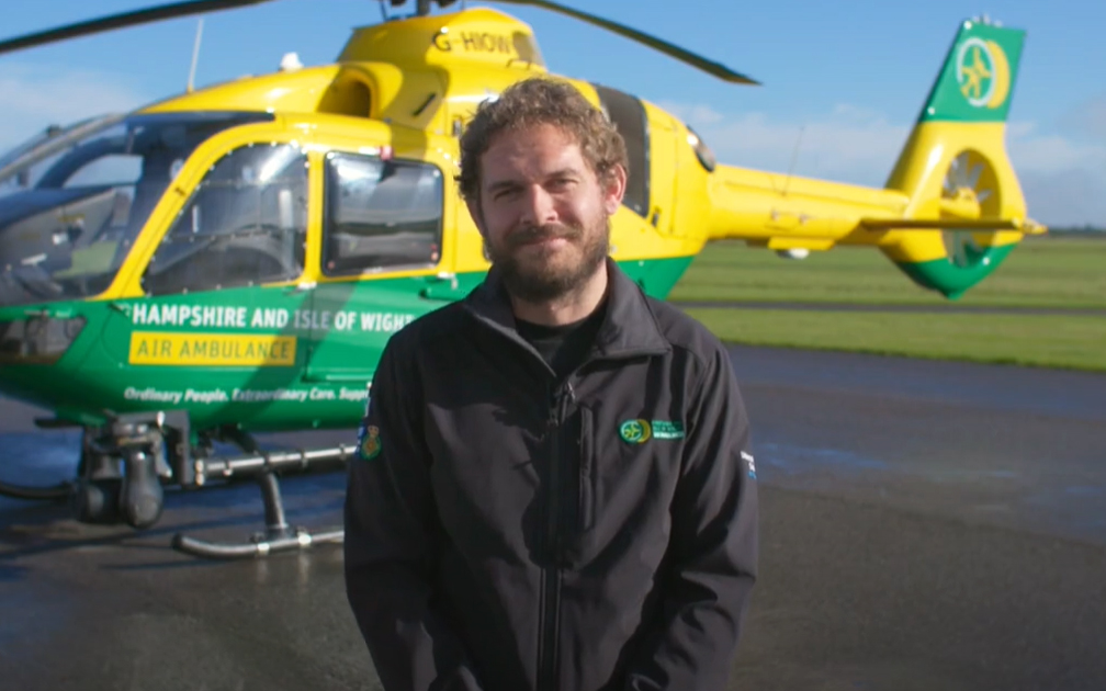 Dr Tom Geliot is stood outside the hangar and in front of the green and yellow helicopter. He is wearing a black jacket and is smiling to camera.