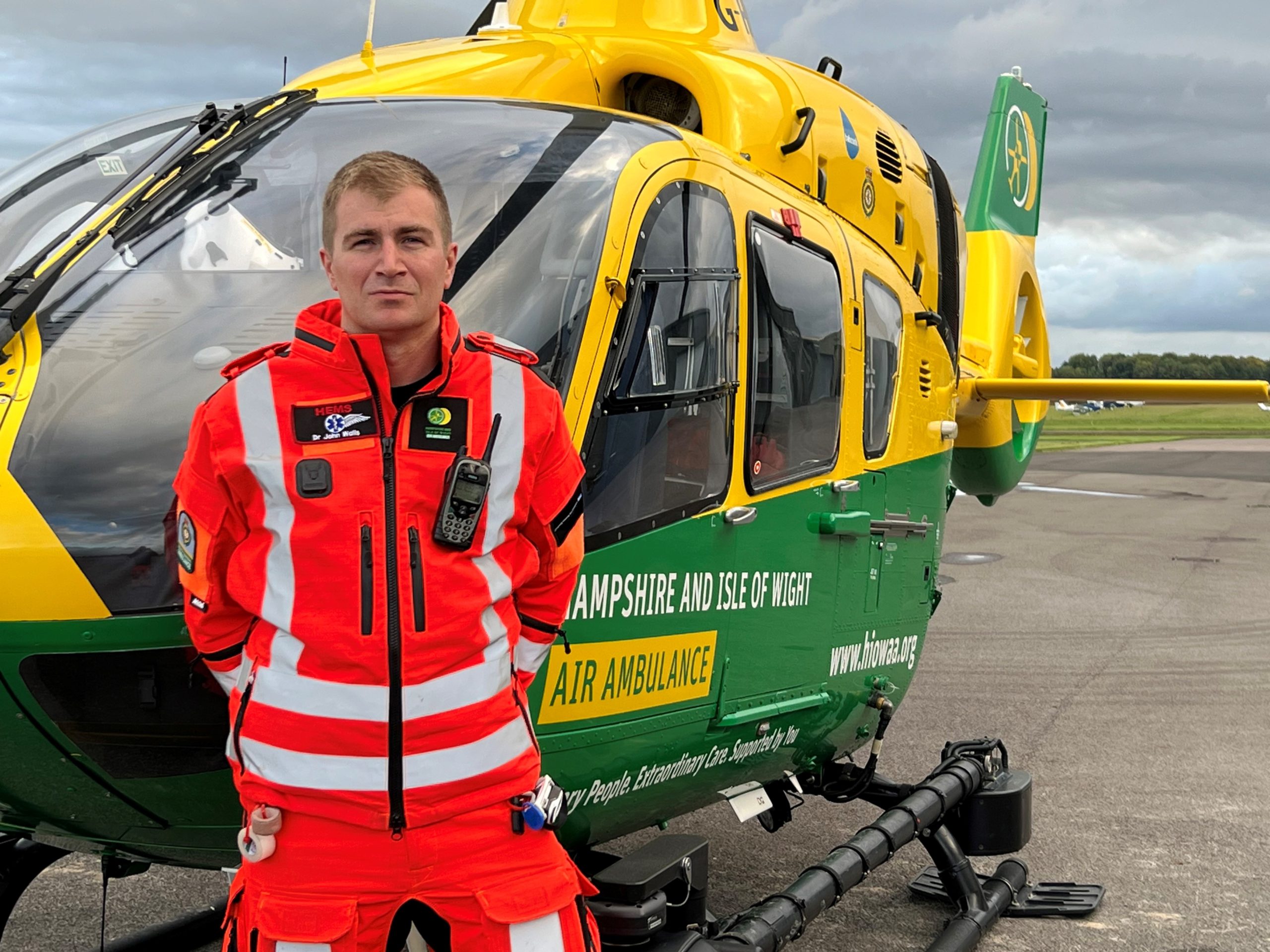 Dr John Walls is stood in front of the helicopter, wearing his flight suit.