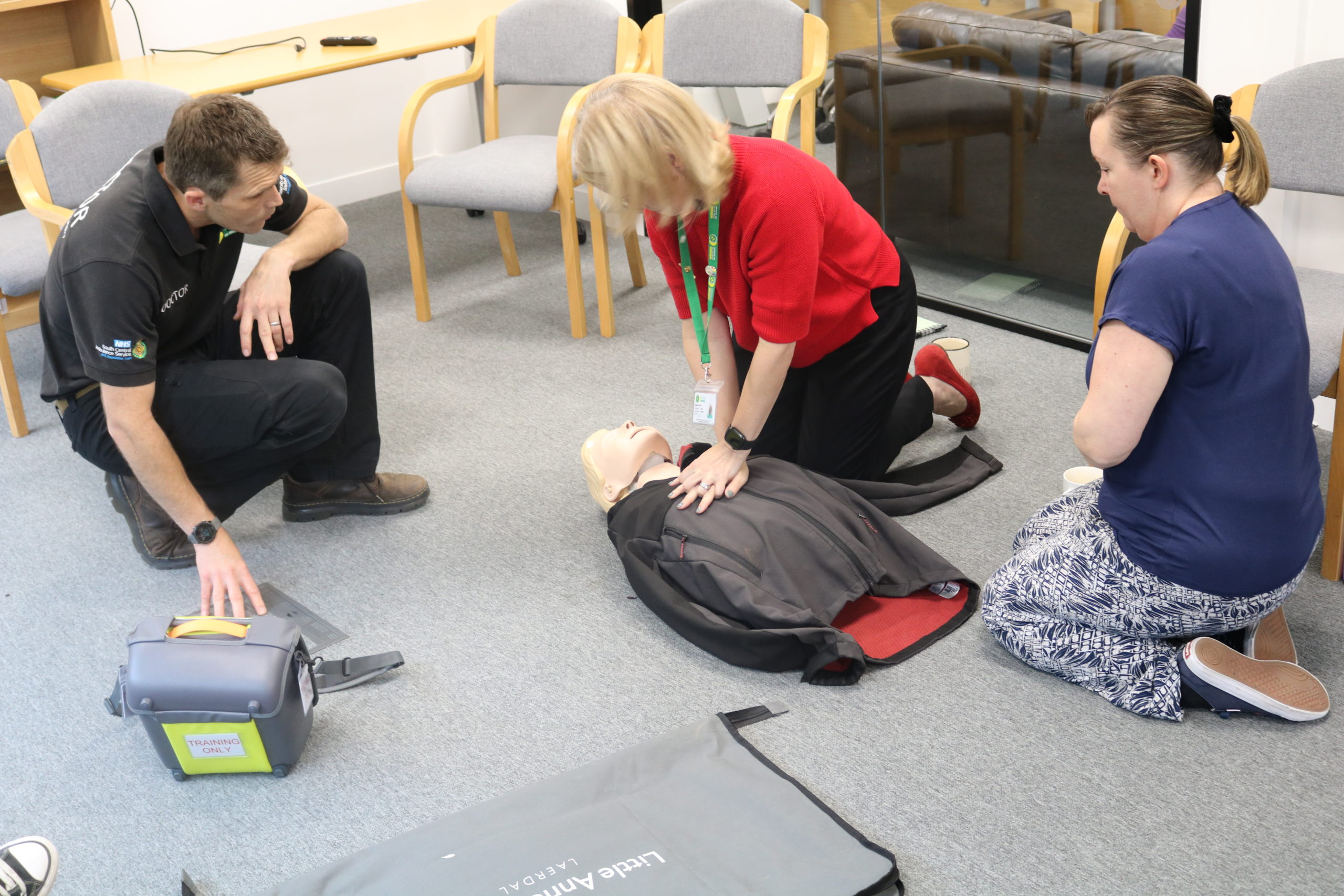 A room of three people. One of them is performing chest compressions on a manikin.