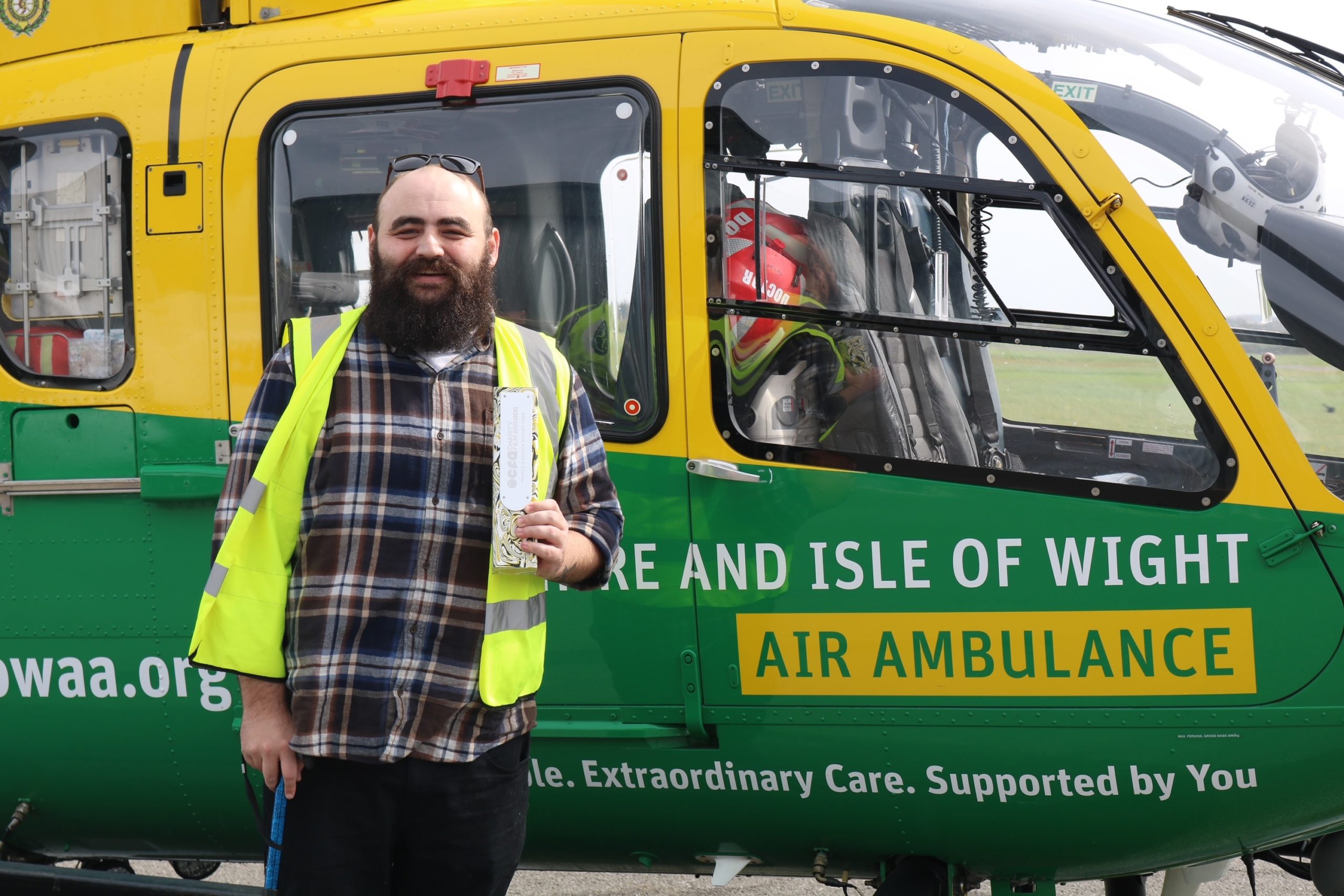 James is stood in front of the helicopter. He is wearing a high-visibility jacket and is smiling to camera.