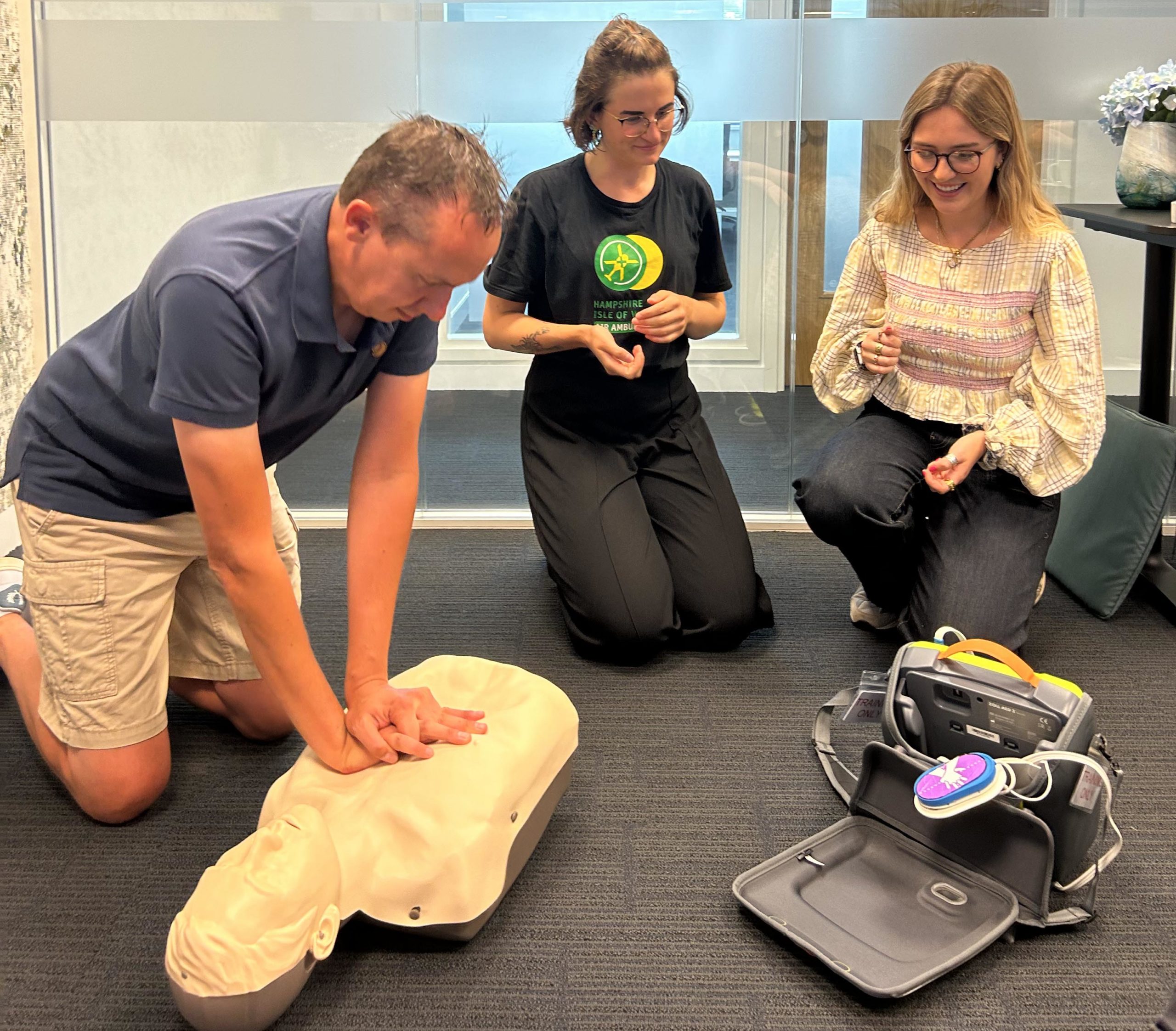 A group of three people kneeling in an office space. One person is performing CPR on a manikin, while the other two are looking at a defibrillator.