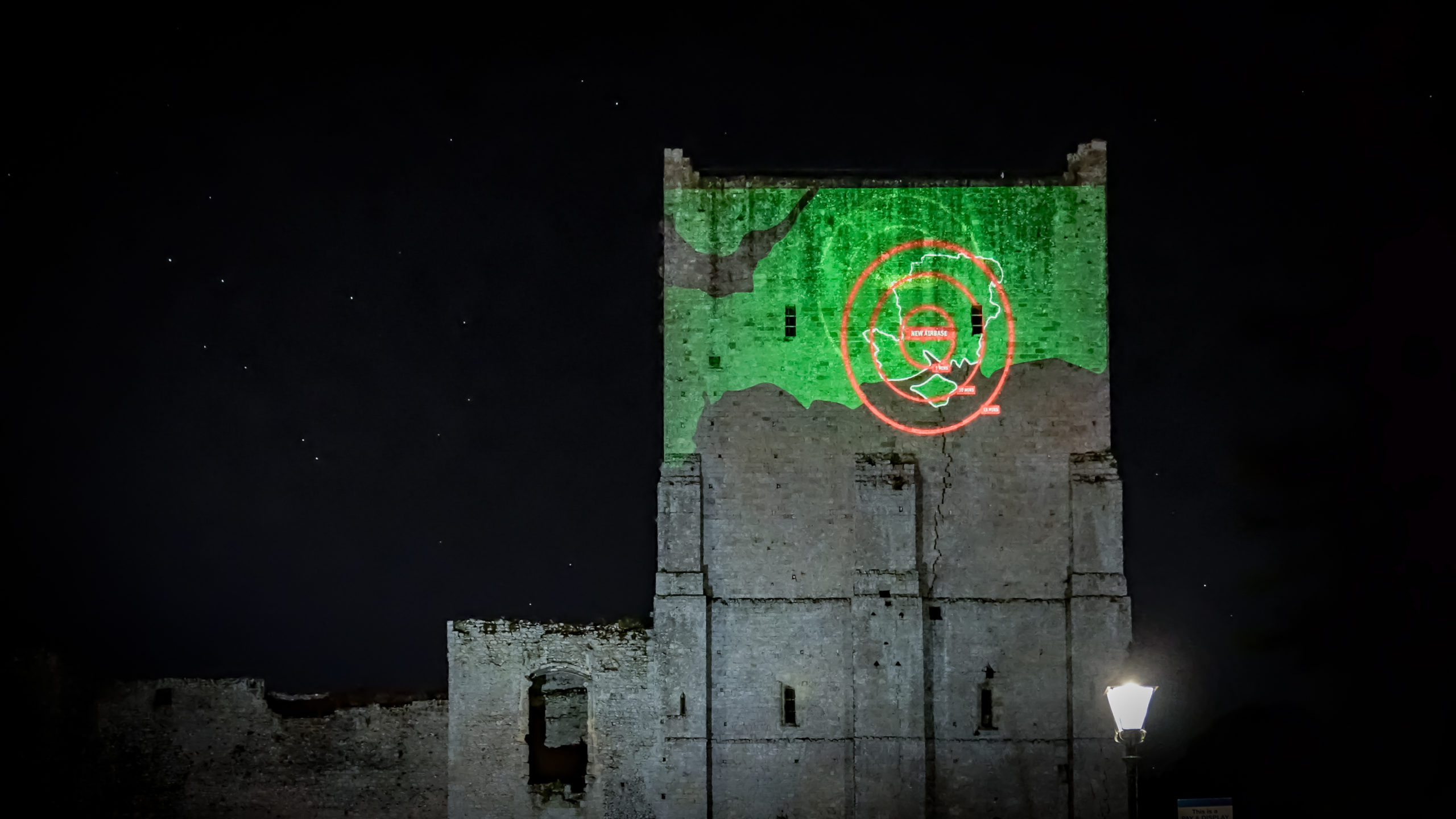 A projection of a map with red circles indicating flight times being displayed on the top of half of Portchester Castle. It is night and there are stars shining behind the castle.