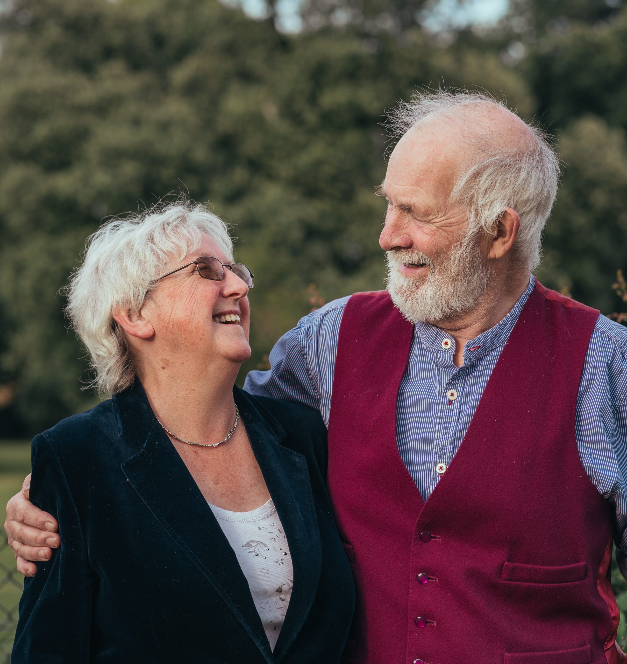 A man and a woman stood side-by-side, looking at one another smiling and laughing. He has his arm around her arm.