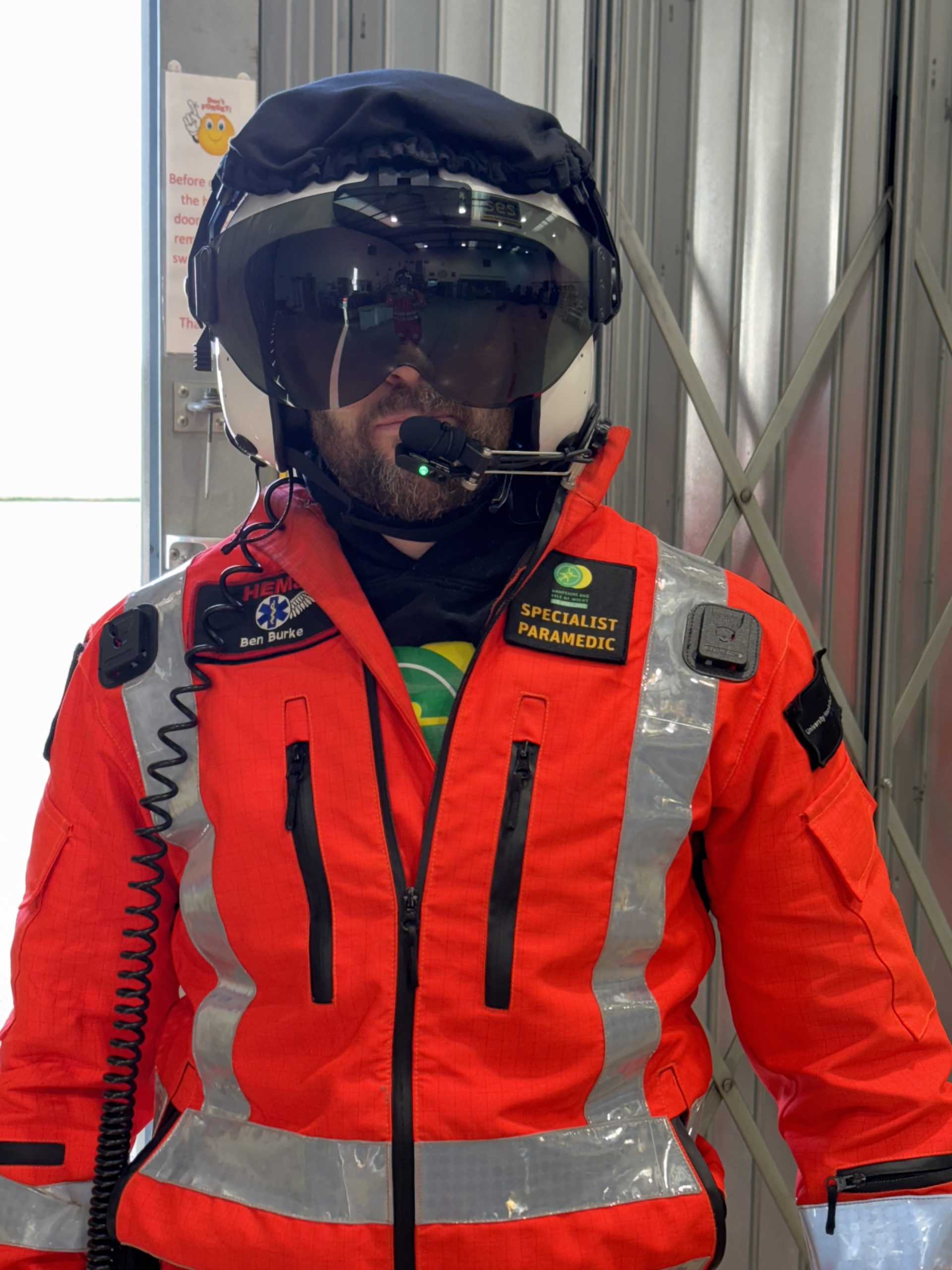 Specialist Paramedic Ben is stood in the airbase hangar in full orange flight suit with his flight helmet on.