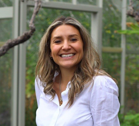 Charity Trustee Lotti Bruce is standing in a garden, smiling to camera. She has shoulder length light brown hair and is wearing a white shirt.