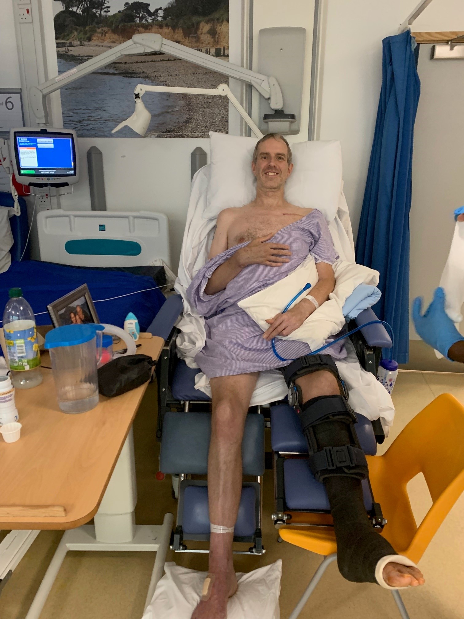 A man in a large chair on a hospital ward. He has his a cast covering his left leg up to and around his knee.