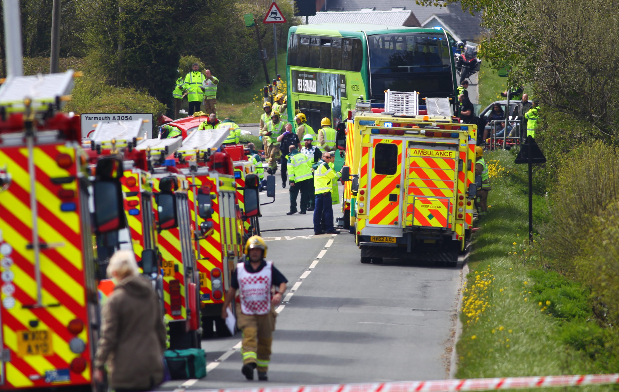 A number of fire engines and ambulances parked on the side of a rural road. There is a green double decker bus at the far end of the picture, which is surrounded by paramedics and fire officers.