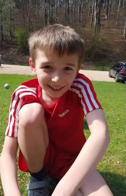 A boy sitting on grass in a woodland area. He is smiling at the camera.