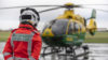 A specialist paramedic in an orange flight suit stood with their back to camera looking at a helicopter in the distance. The helicopter is on the ground preparing for take off.