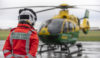 A specialist paramedic in an orange flight suit stood with their back to camera looking at a helicopter in the distance. The helicopter is on the ground preparing for take off.