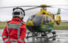 A specialist paramedic in an orange flight suit stood with their back to camera looking at a helicopter in the distance. The helicopter is on the ground preparing for take off.